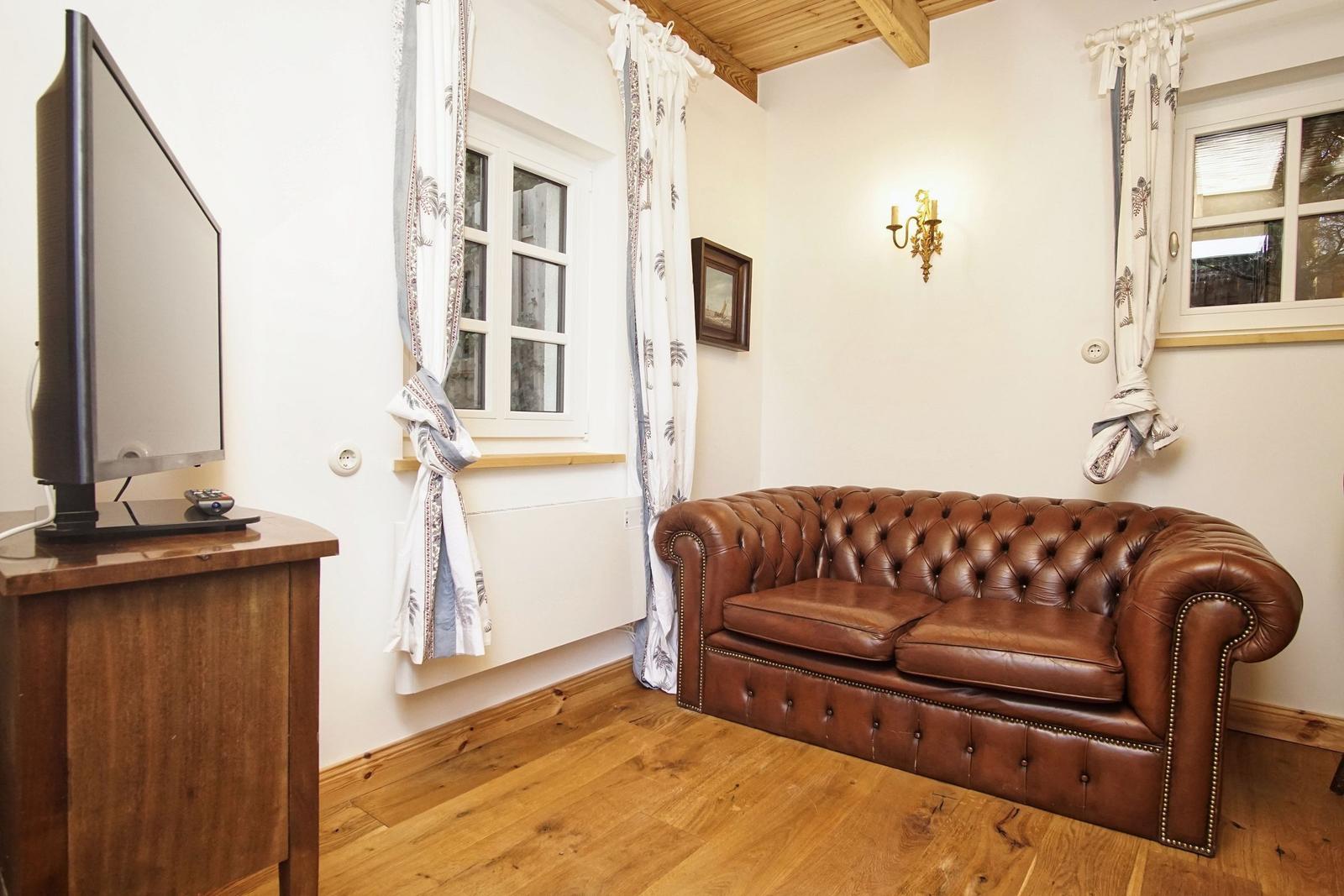 Ein Wohnzimmer mit braunem Ledersofa, Holzboden und TV auf Holzkonsole.