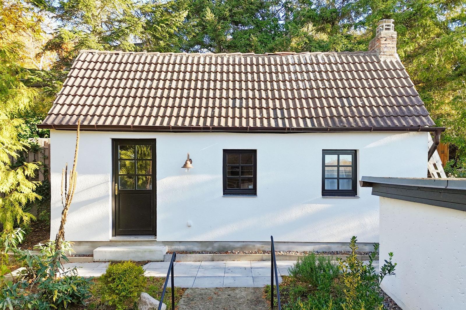 Wohnhaus mit weißer Fassade, schwarzem Eingang und zwei Fenstern, umgeben von Grün.