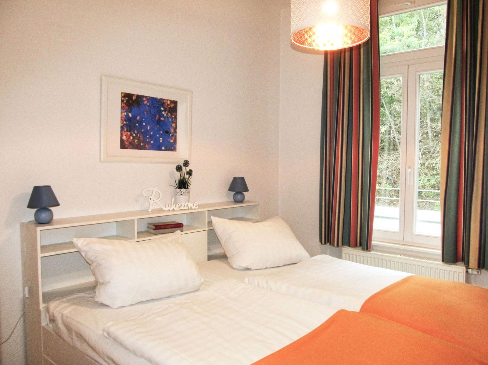 Zimmer mit Doppelbett, Fenster, Vorhängen und Deckenleuchte.