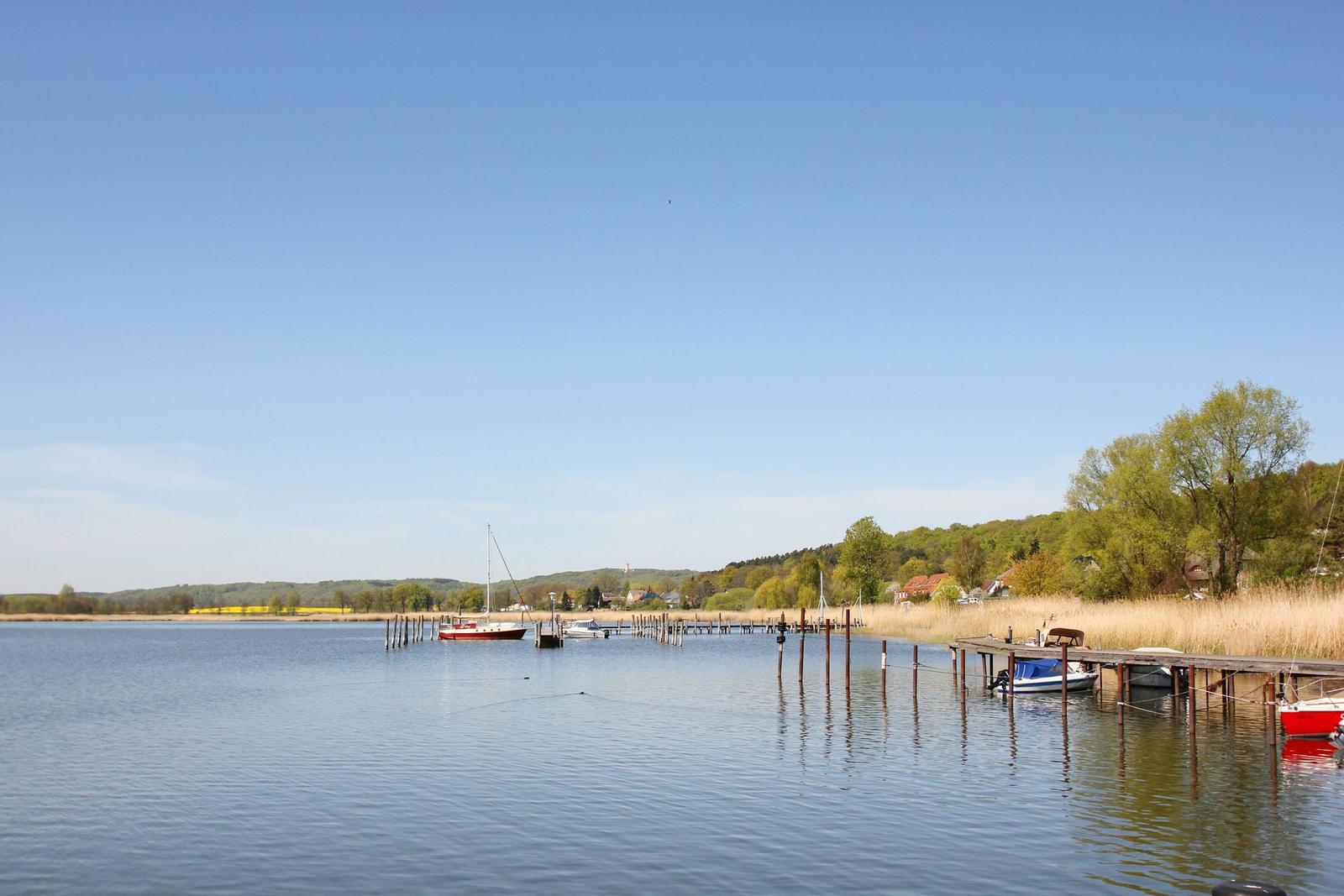 Stille See mit Booten und Holzstegen. Grün bewachsener Uferbereich mit Häusern im Hintergrund.