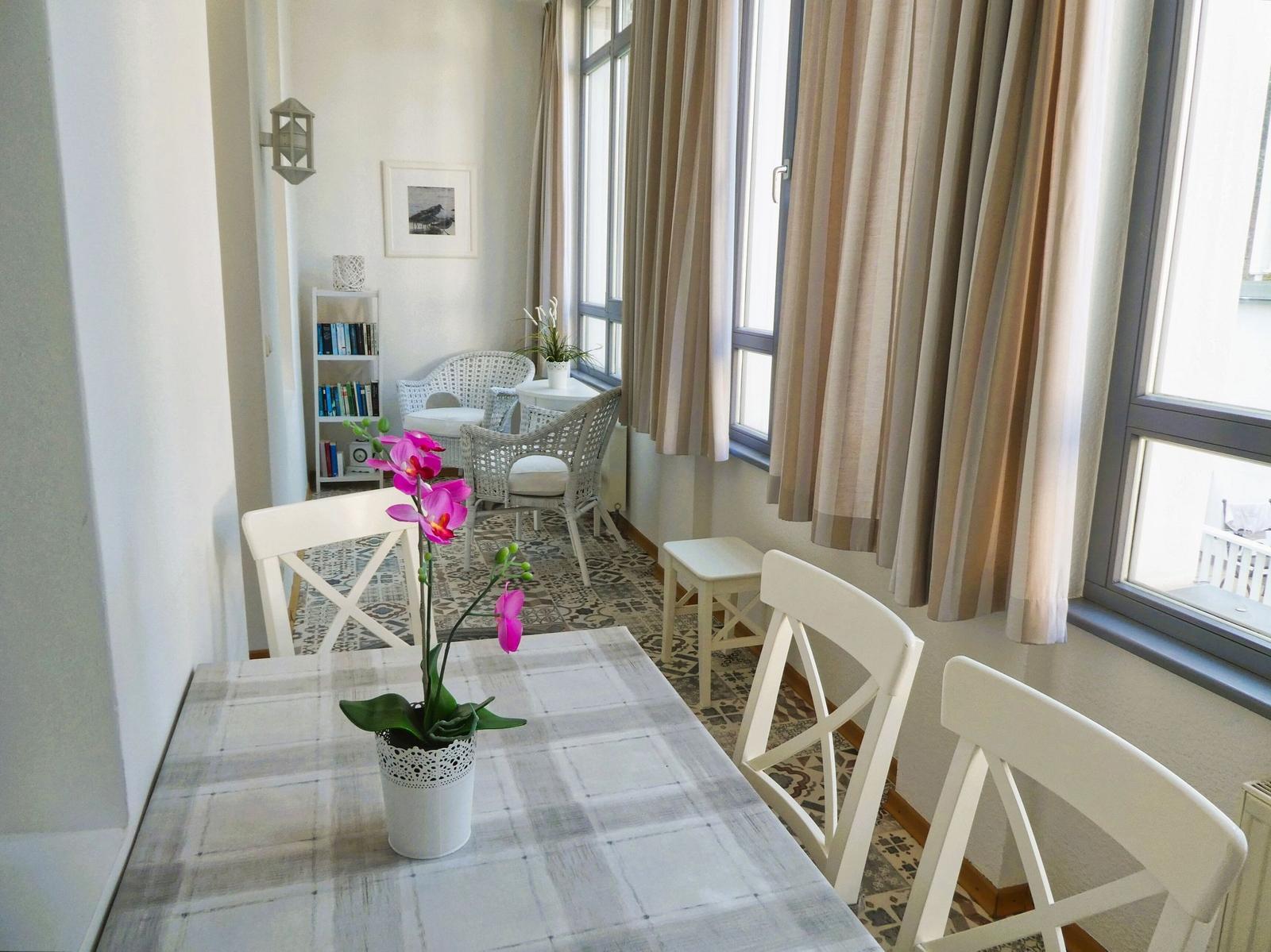 Essbereich mit Tisch, Stühlen und Blume. Im Hintergrund Sitzbereich mit Bücherregal.