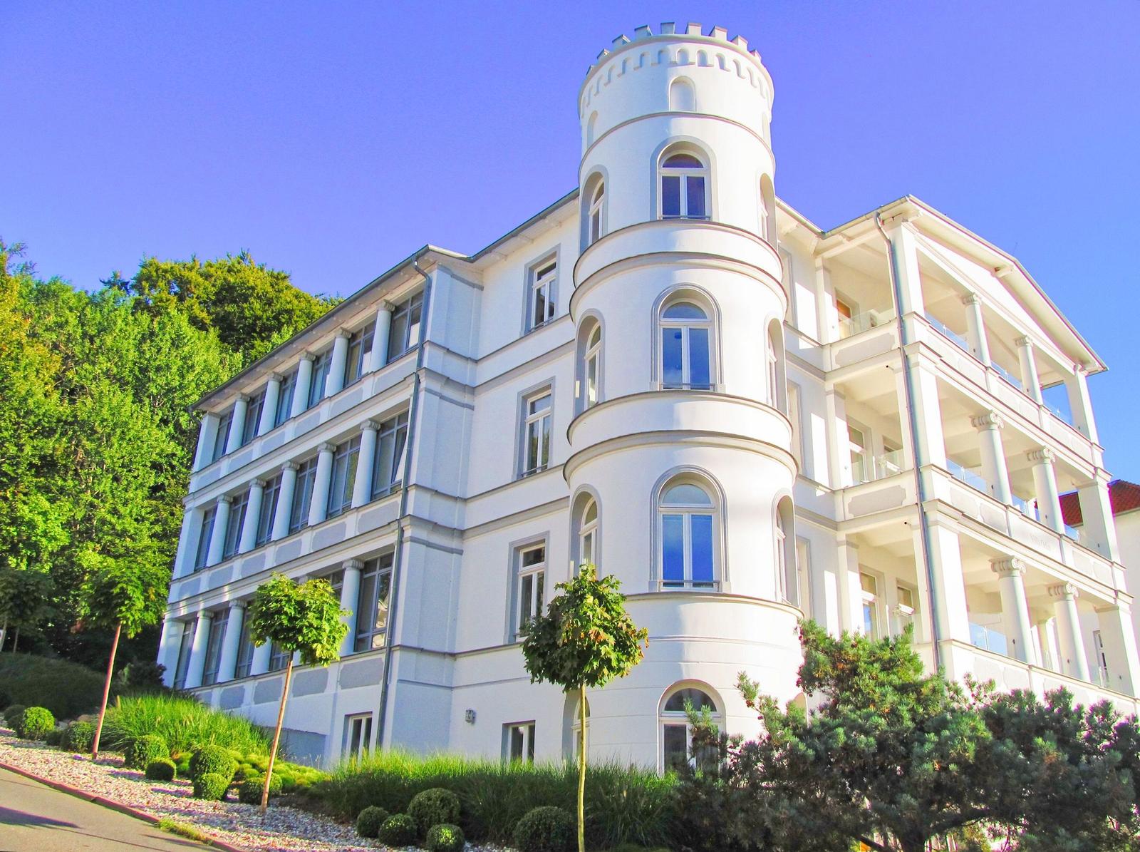 Weißes Gebäude mit Turm und Balkonen, umgeben von Grünflächen und Bäumen.