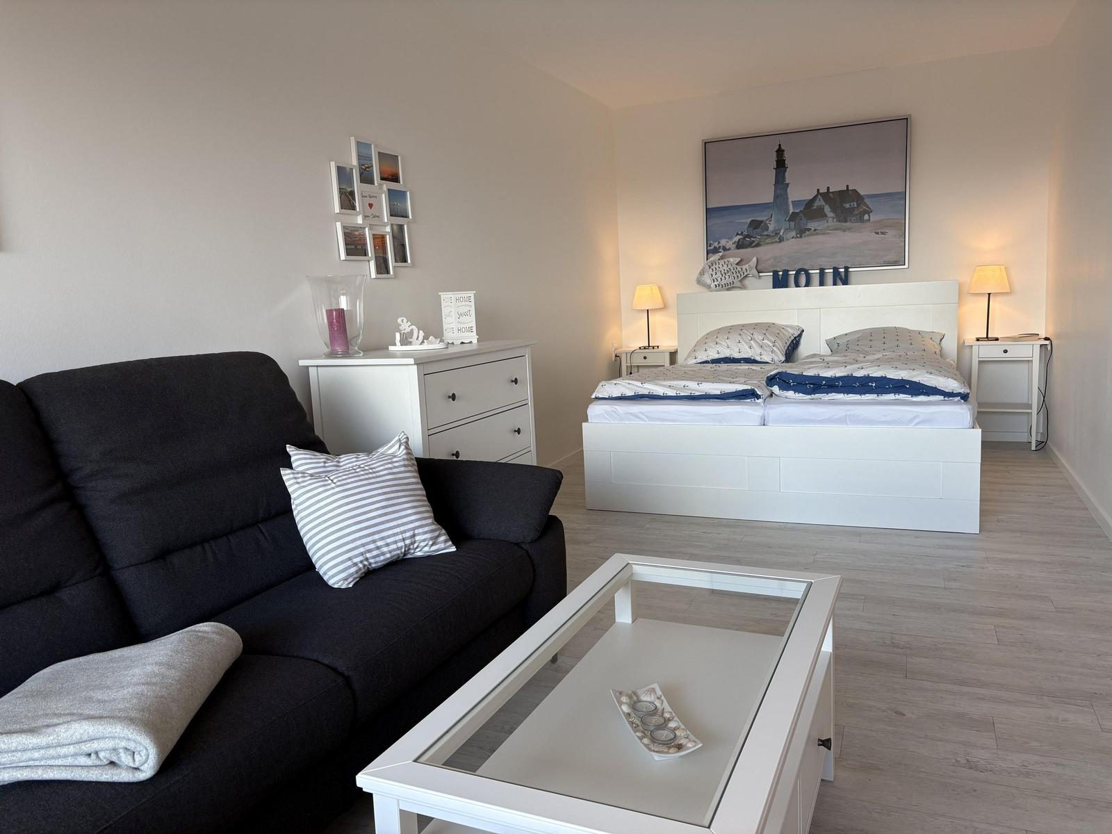 Schlafzimmer mit Sofa, Bett und Tisch. Wandbild zeigt Leuchtturm.