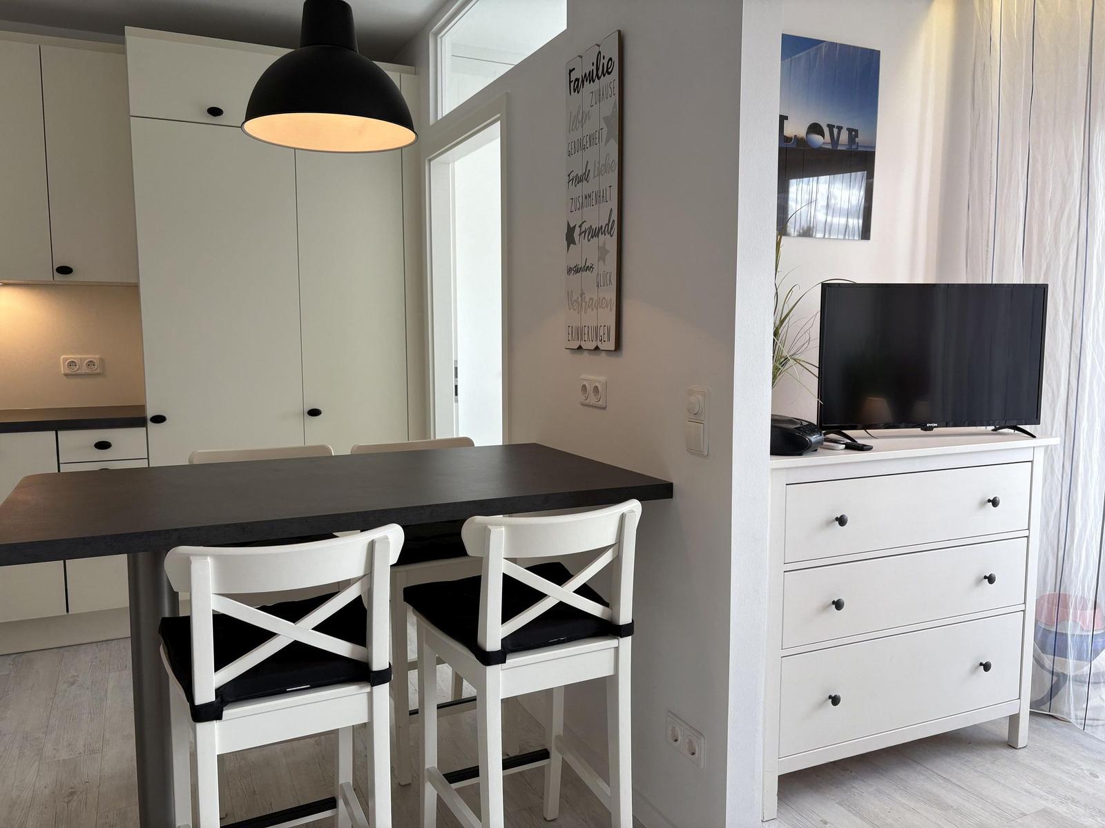 Essbereich mit Tisch, Stühlen und Küchenzeile. Fernseher auf Schrank.