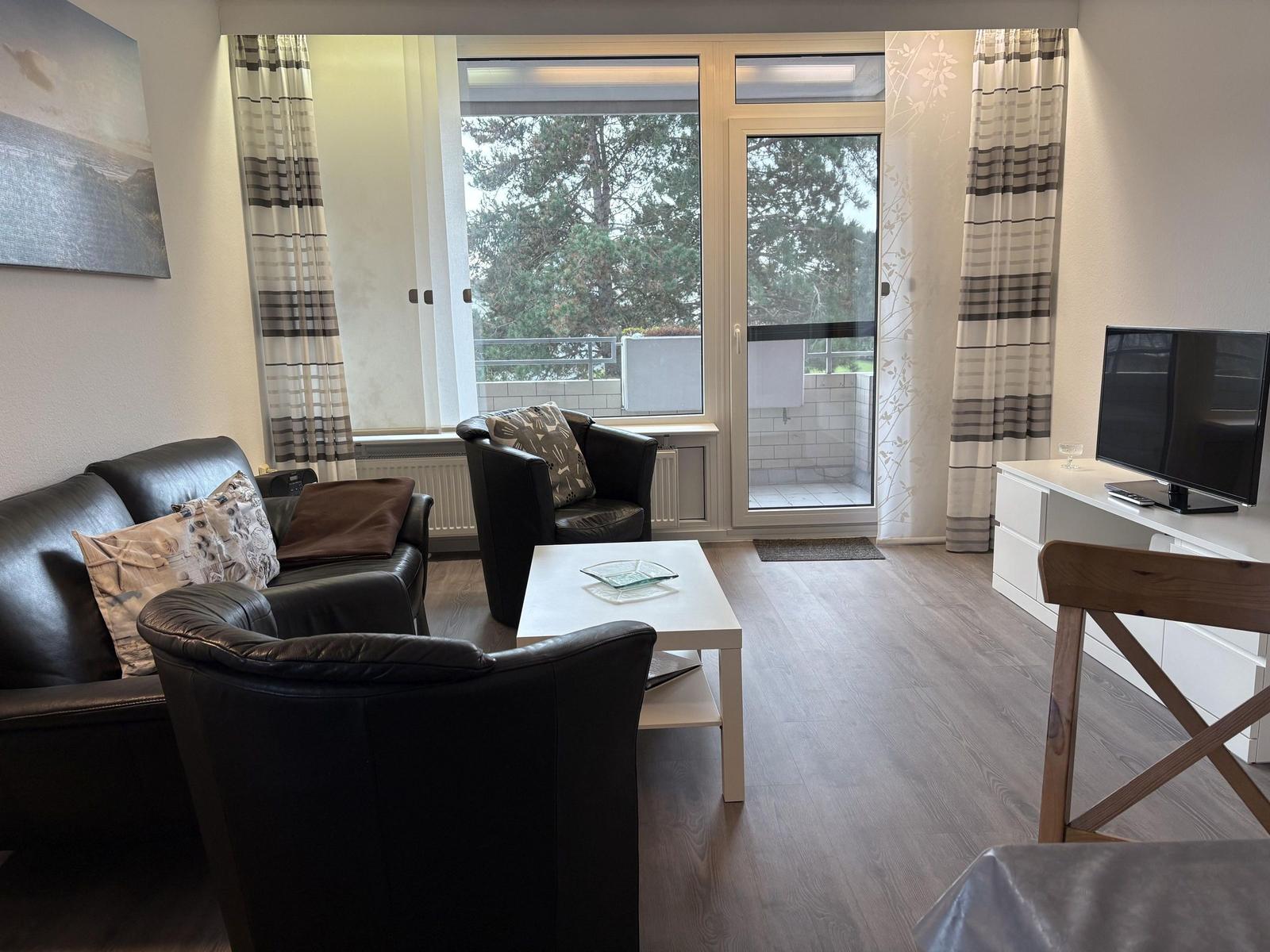 Wohnzimmer mit Ledersofa, Sessel und TV. Balkon mit Blick auf Bäume.