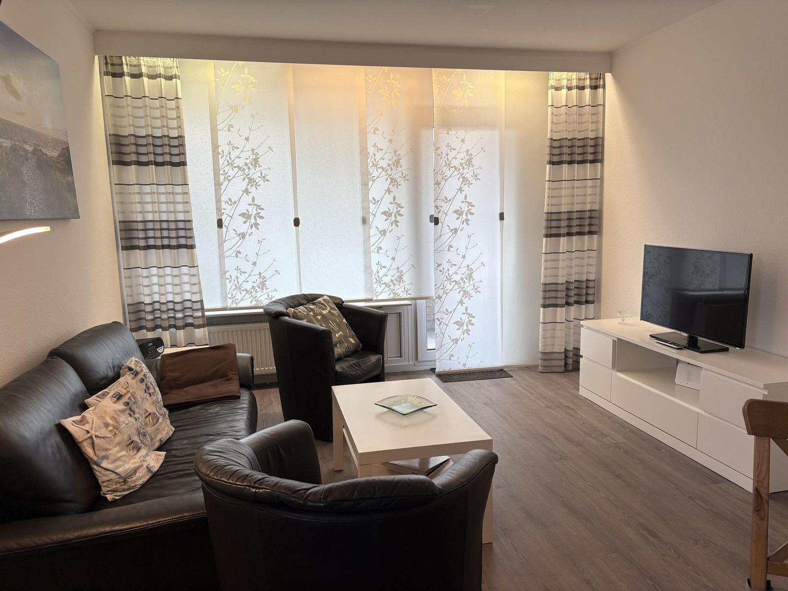 Wohnzimmer mit Sofa, Sesseln, TV und Fenster mit Vorhängen.