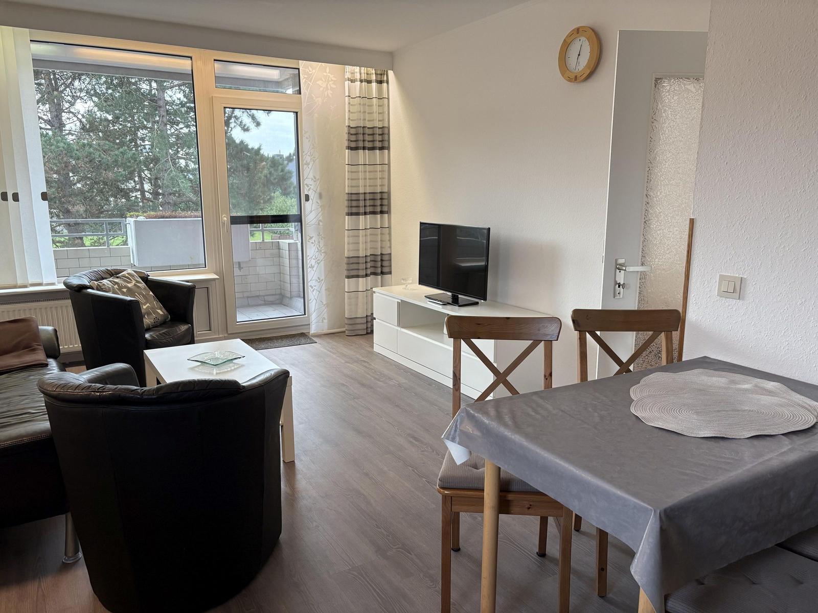 Wohn- und Essbereich mit Fernseher, Tisch und Stühlen. Balkon mit Blick auf Bäume.