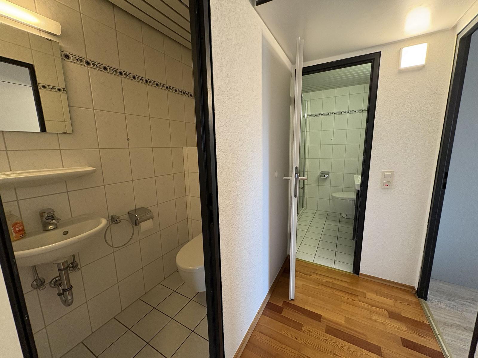 Badezimmer mit Waschbecken, Spiegel und Toilette. Durchgang zur Dusche mit WC.