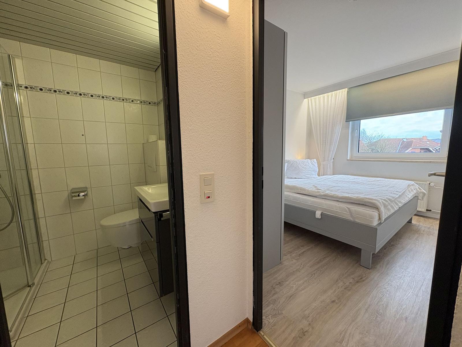 Schlafzimmer mit Bett und Fenster. Nebenraum ist Badezimmer mit Dusche.
