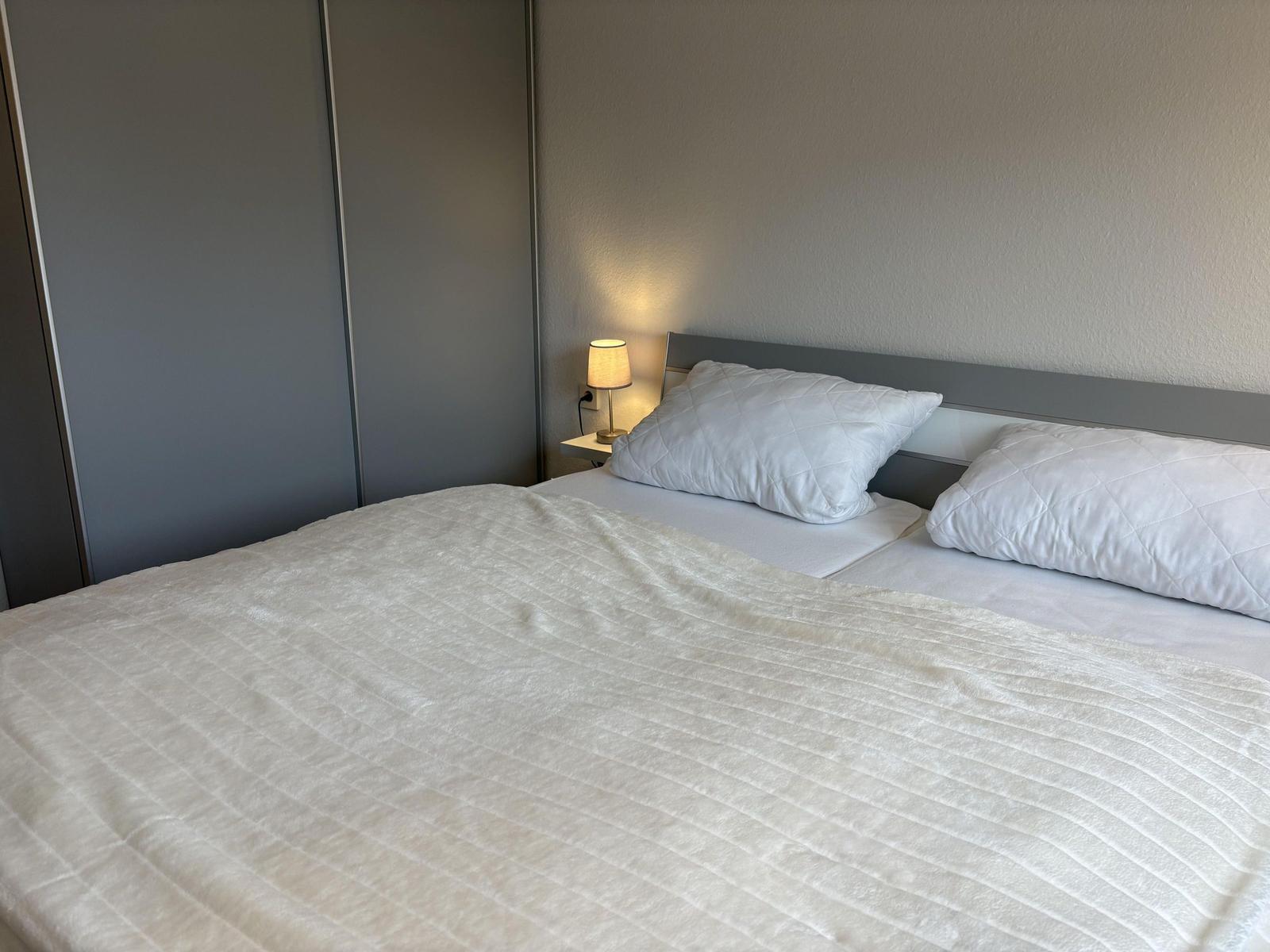 Schlafzimmer mit Bett, Kissen und Nachttischlampe. Hintergrund: Schrank und Wand.