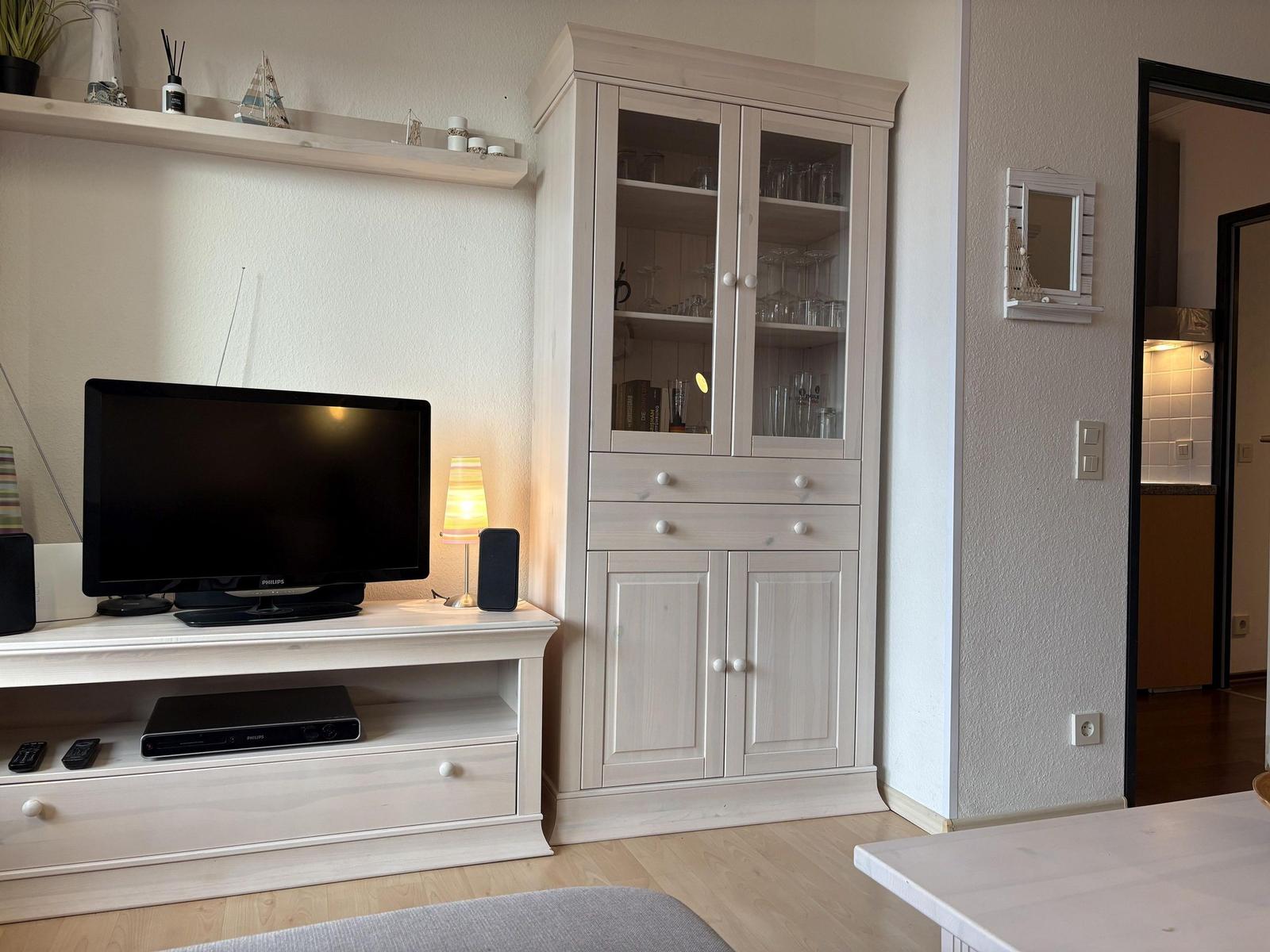 TV-Möbel mit Fernseher und Glaswand-Schrank. Küche im Hintergrund sichtbar.