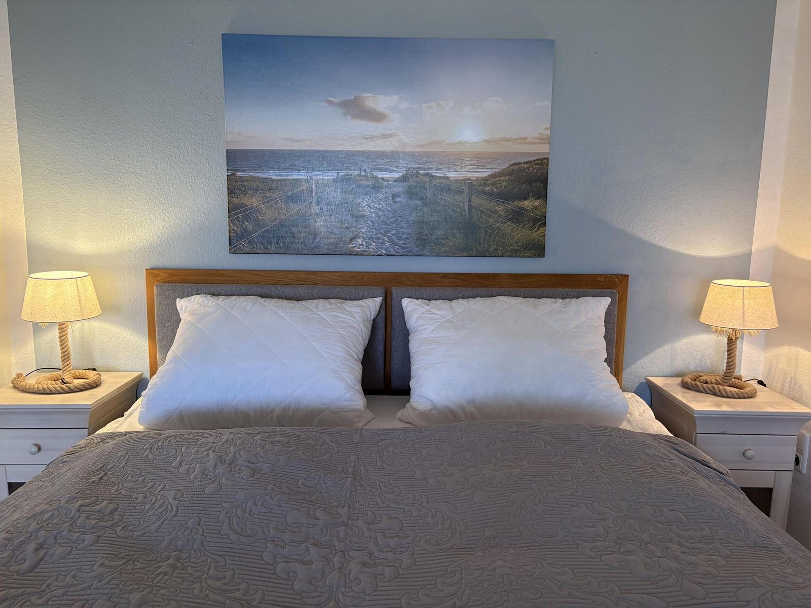 Doppelbett mit Kopfkissen, Nachttische und Wandbild mit Strandmotiv.