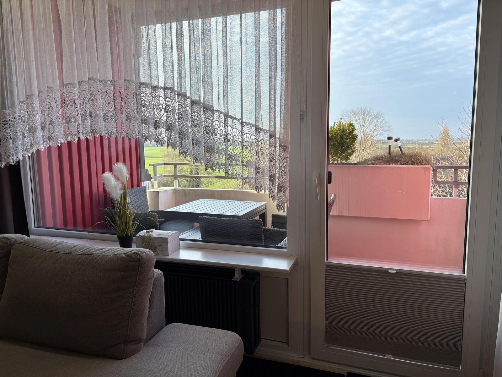 Durch das Fenster ist eine Terrasse mit Tisch und Stühlen zu sehen. Vor dem Fenster steht ein Sofa.