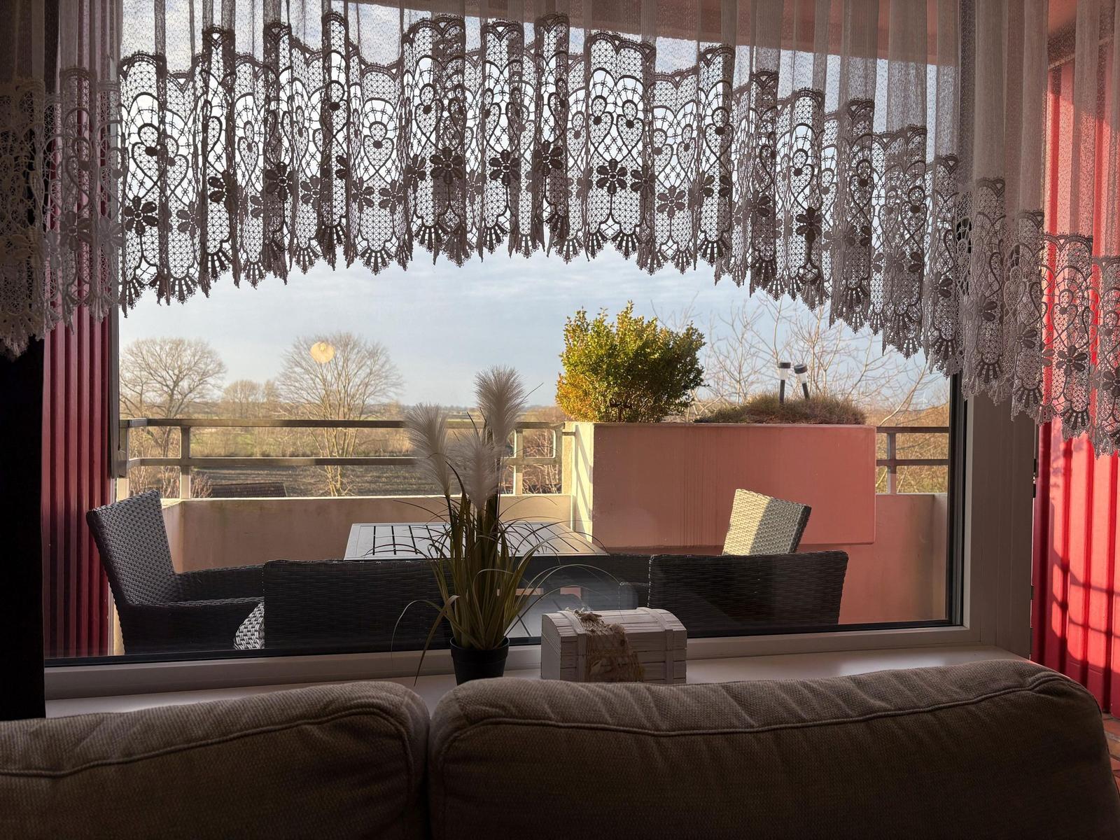 Durch das Fenster sieht man einen Balkon mit Sitzgelegenheiten und Blick auf die Landschaft.