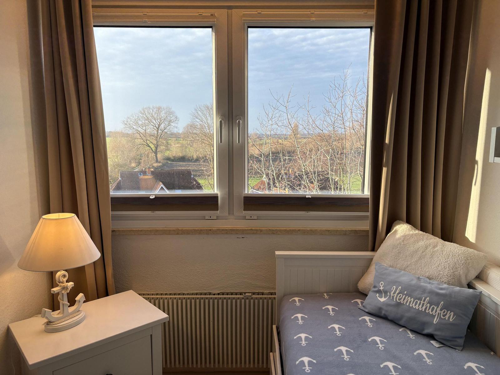 Schlafzimmer mit Blick auf Felder und Bäume. Bett mit Ankern-Motiv und Nachttisch mit Lampe.