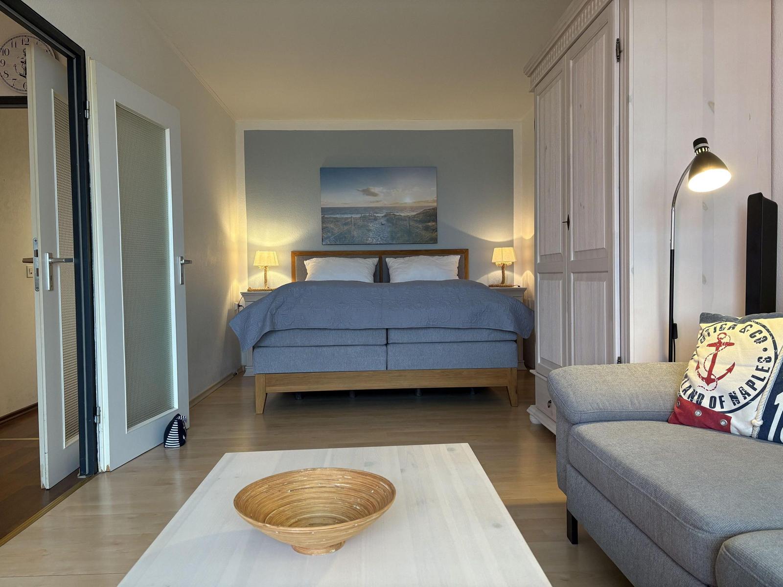 Schlafzimmer mit Bett, Couch und Wandbild. Helle Holzböden und weiße Möbel.