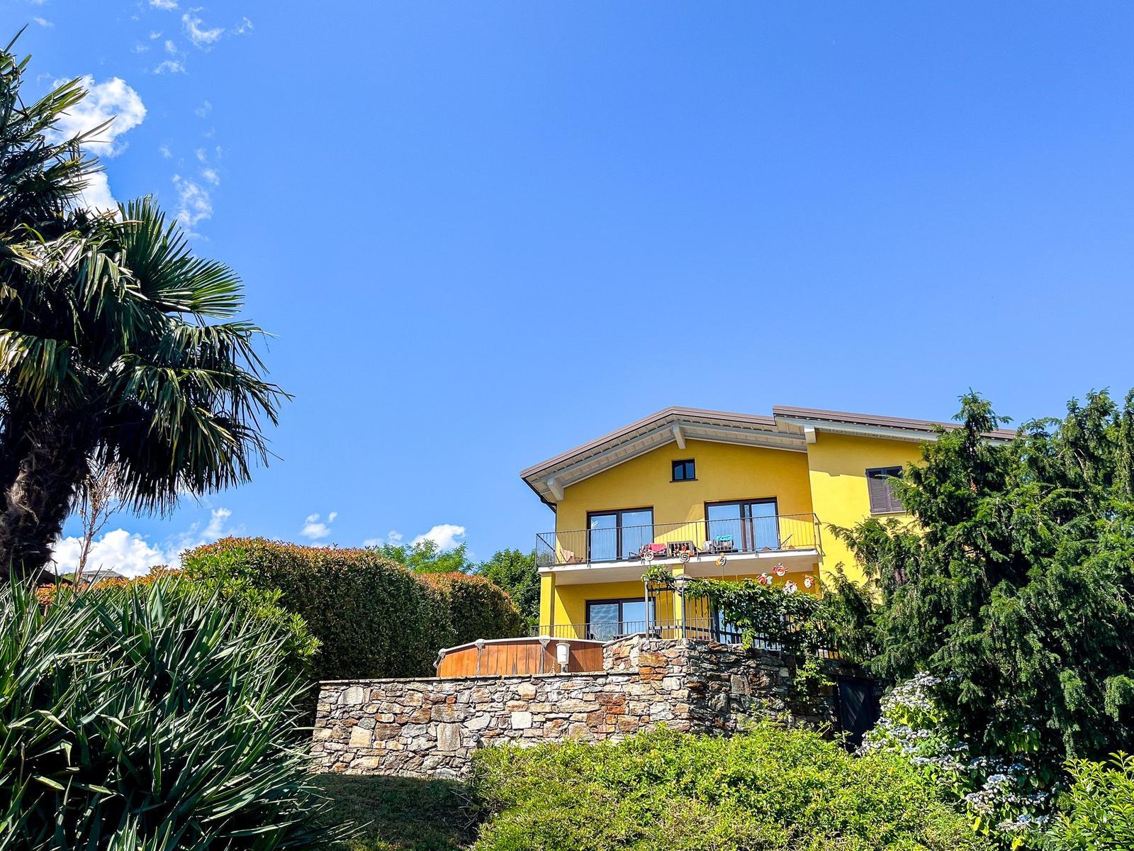 Yellow house with balcony, surrounded by greenery and stone walls under blue sky.