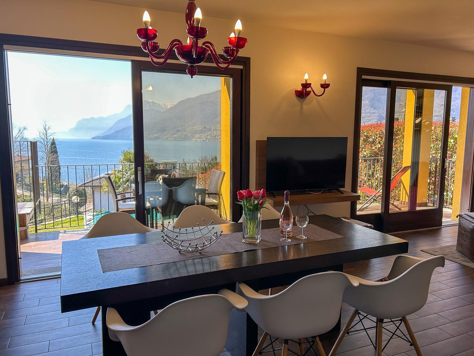 Dining room with lake and mountain view. Table, chairs, TV, and red ceiling lights.