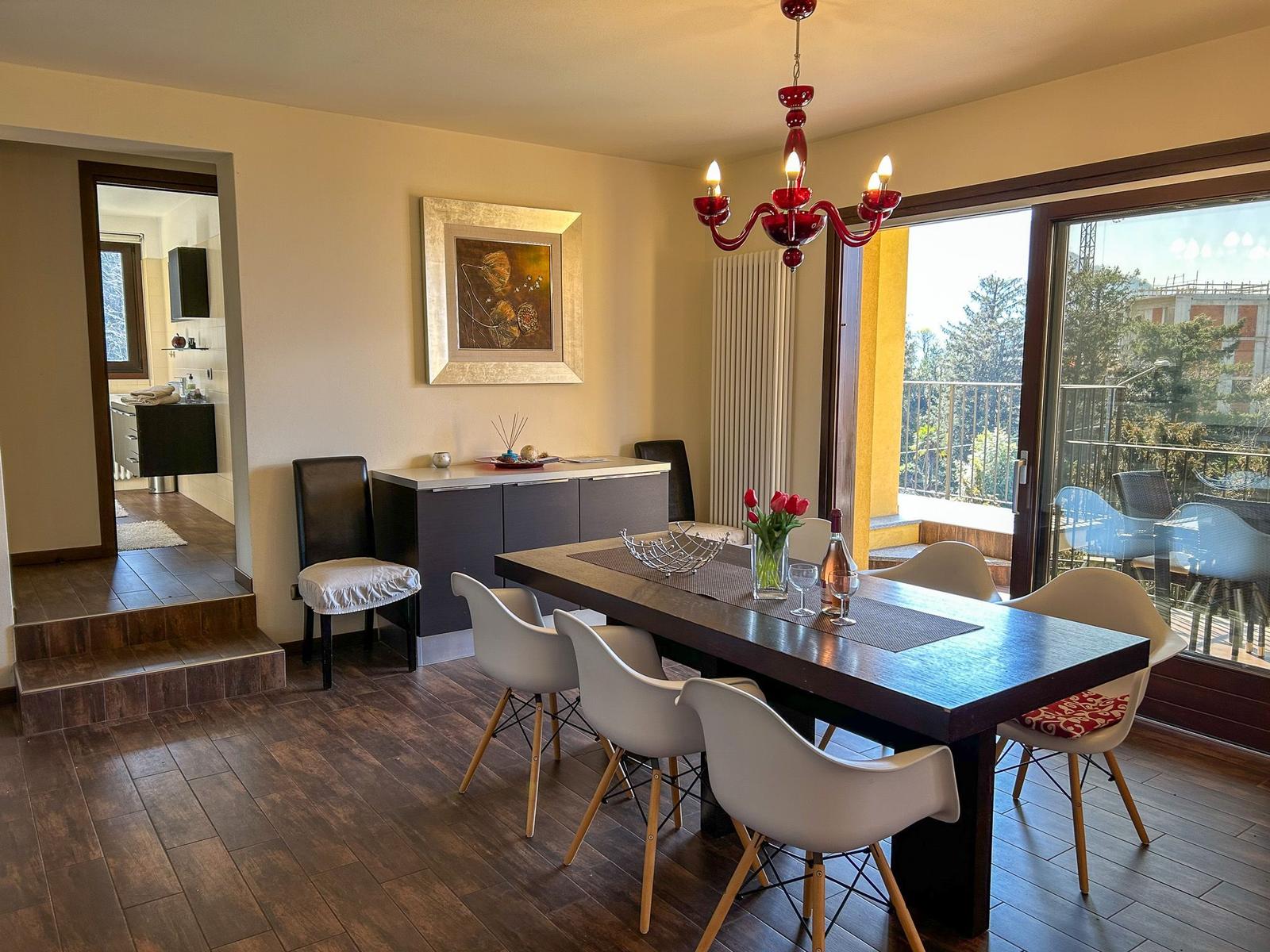 Dining room with table, chairs, and balcony view. Red chandelier and wall art.