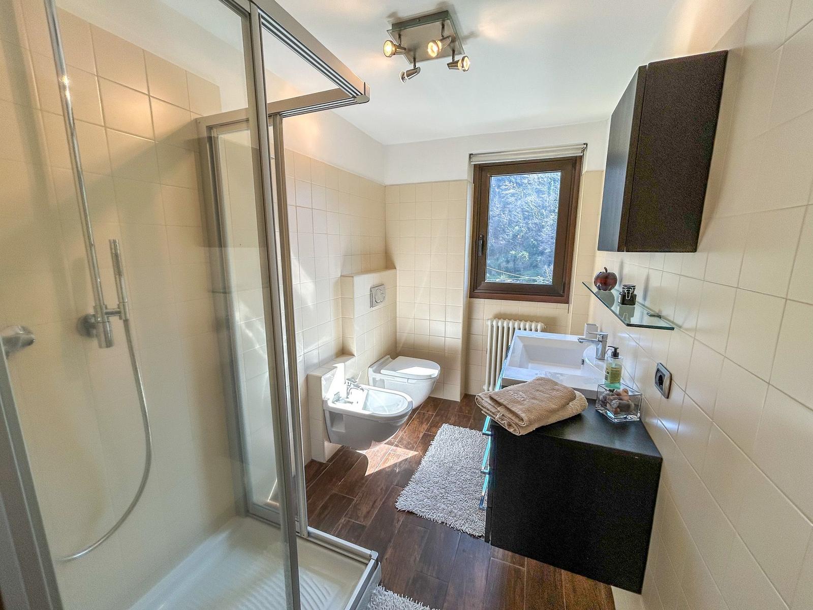 Bathroom with shower, toilet, and sink. Window with view of forest.