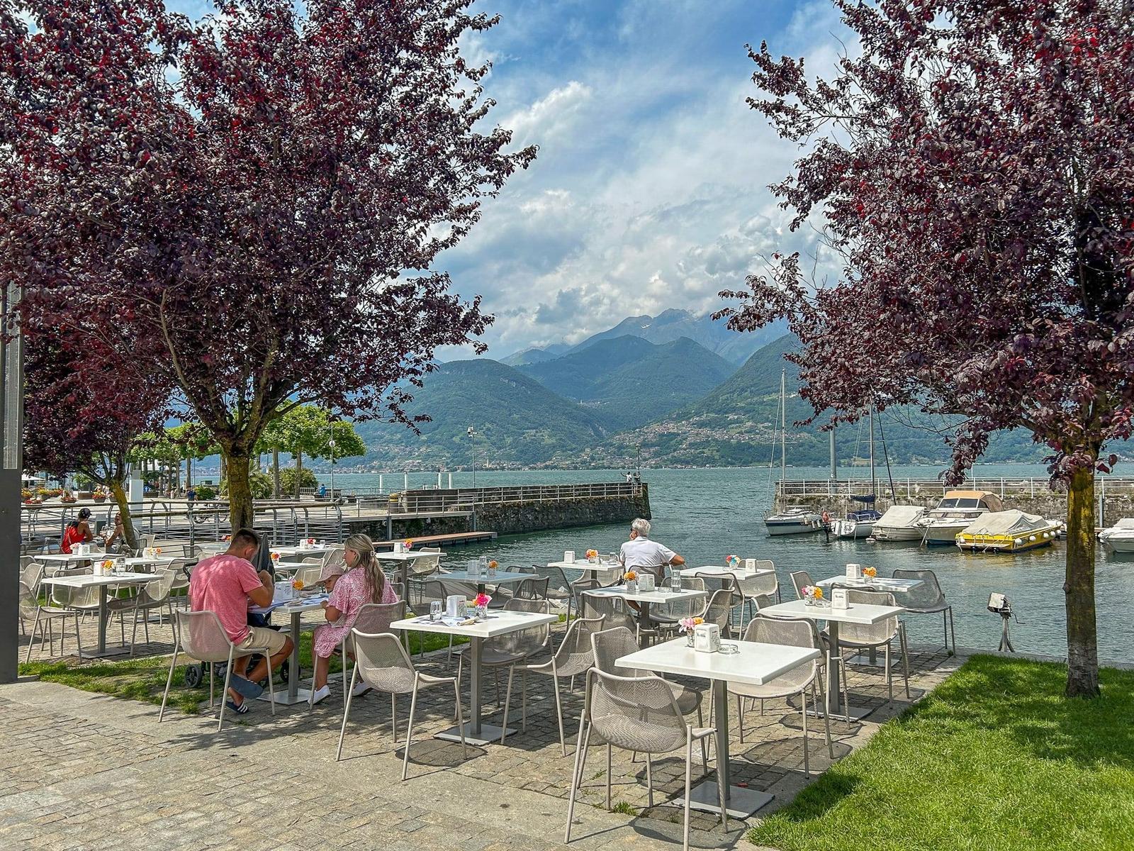 Terrace with tables and chairs by the lake, boats, and mountains in the background.