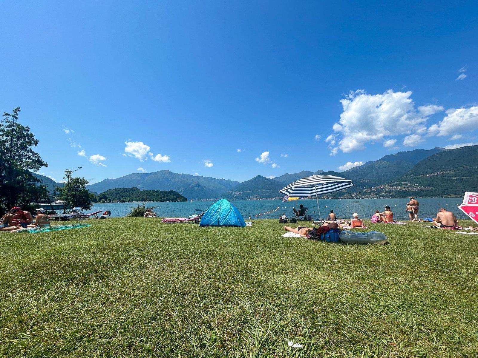 Green shore with people, tent, and beach chairs by the lake.
