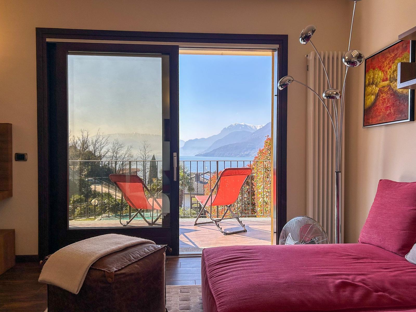 Room with balcony, view of lake and mountains, red sun loungers, bed with pillows.