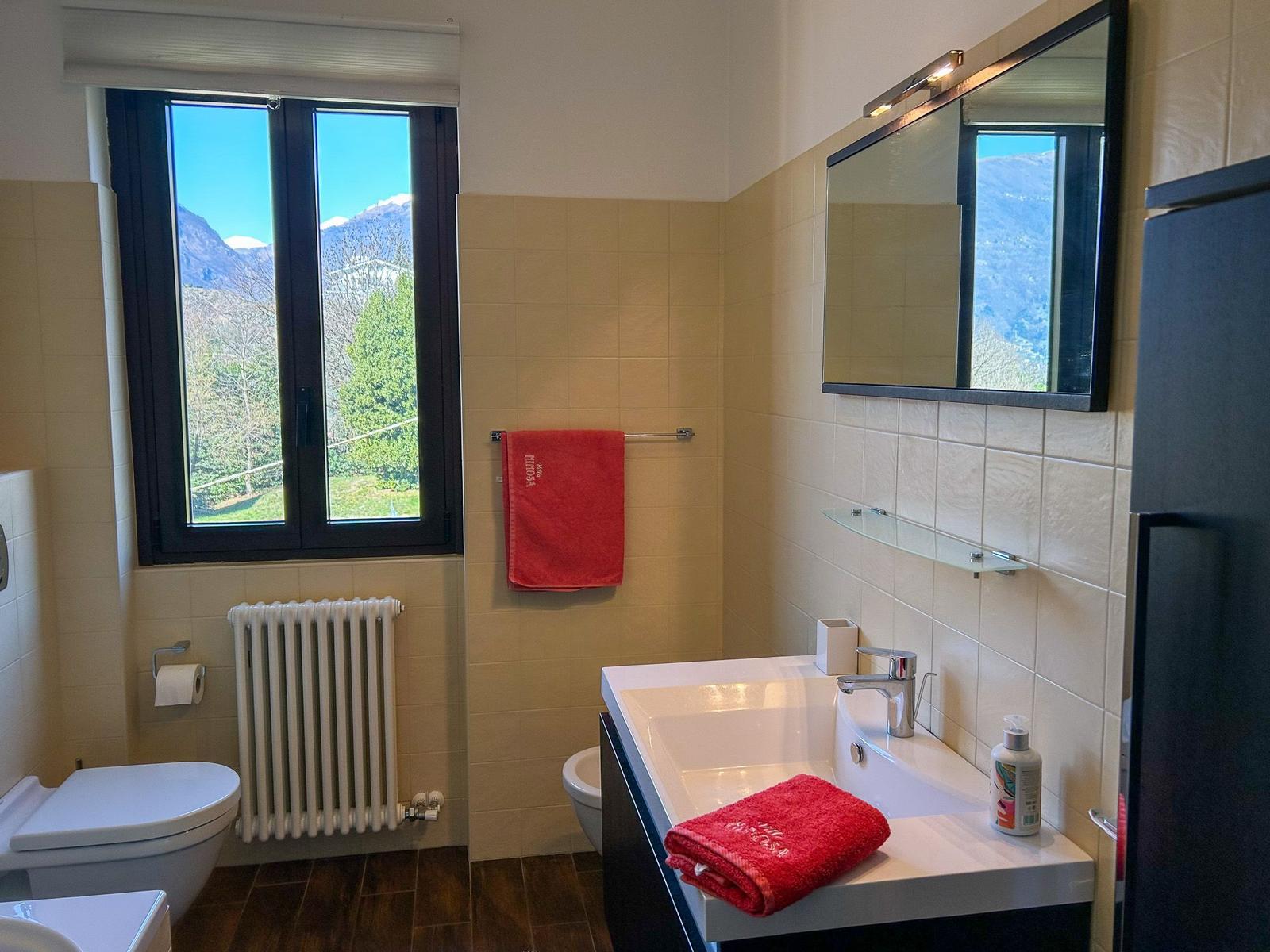 Badezimmer mit Fensterblick auf Berge, Waschbecken, Spiegel und rote Handtücher.