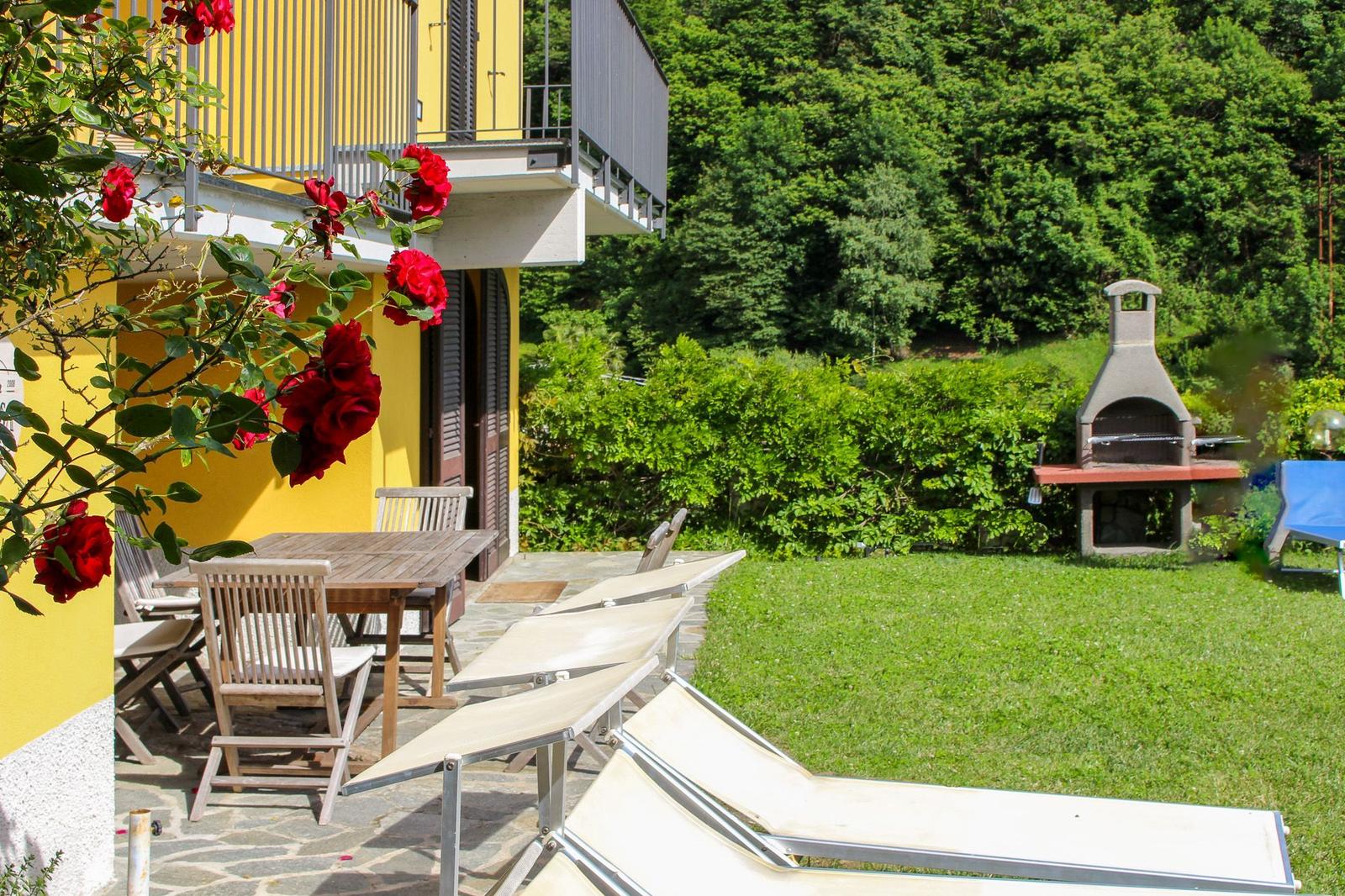 Garten mit Liegestühlen, Grill und Tisch. Gelbes Haus mit Balkon und roten Rosen.