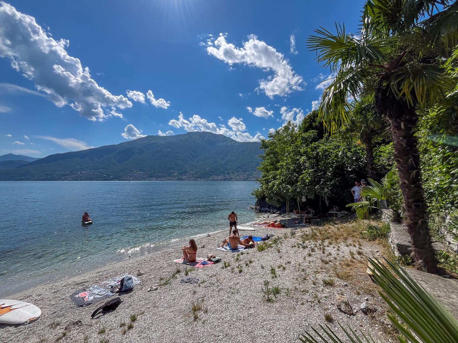 Strand mit Steinen, Palmen und Menschen am Seeufer unter blauem Himmel.