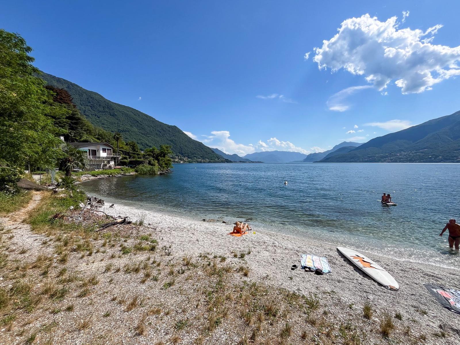 Strand mit Kies, See, Bergen und Sonnenbadenden.