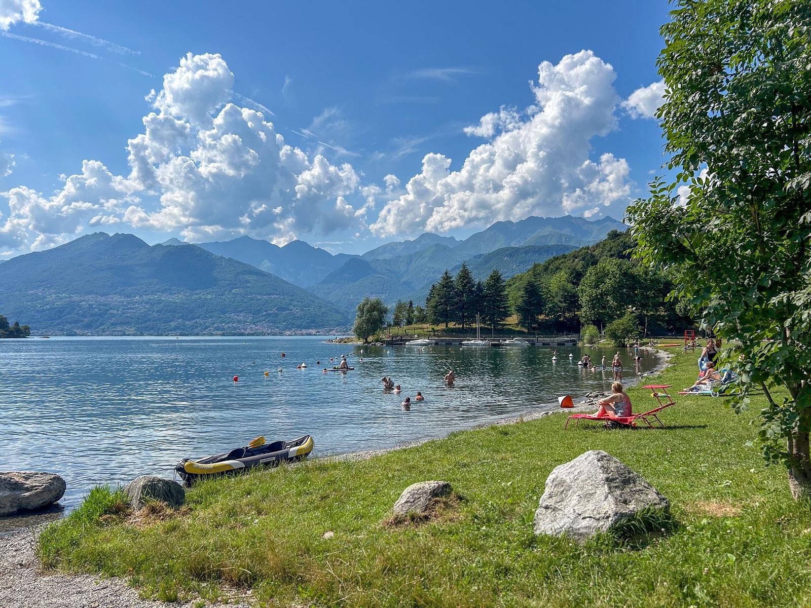 Lakeside with people swimming and relaxing on grass near mountains.