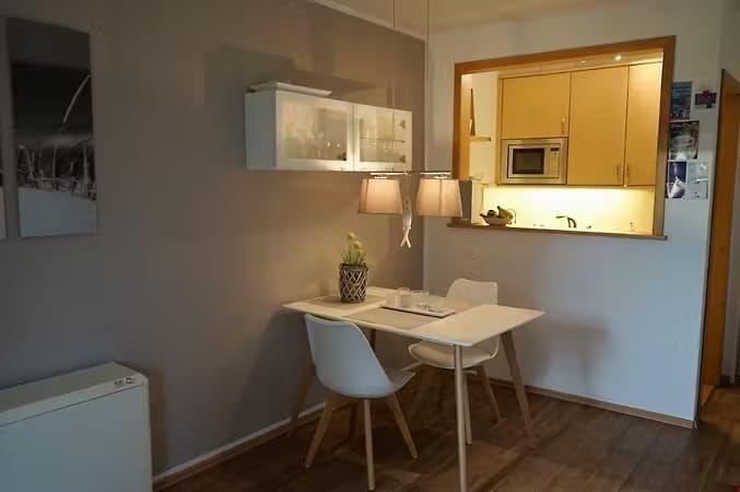 Küche mit Essbereich, weiße Tische und Stühle, Holzboden und hängende Schränke.