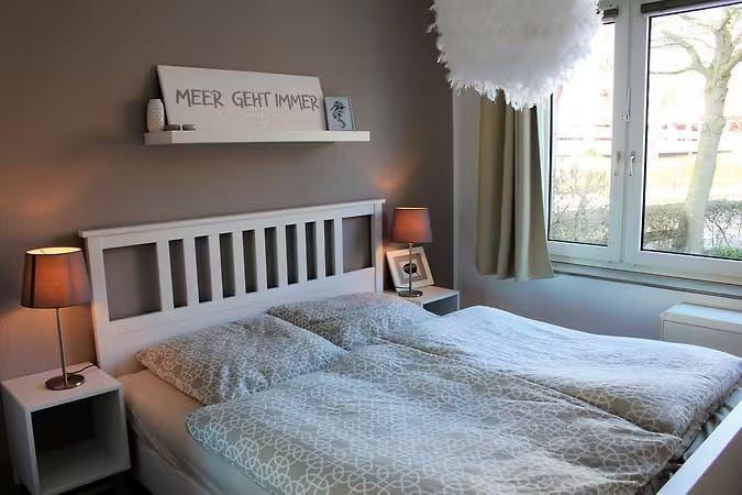 Schlafzimmer mit Bett, Nachttisch und Fenster. Wandregal mit Schriftzug.