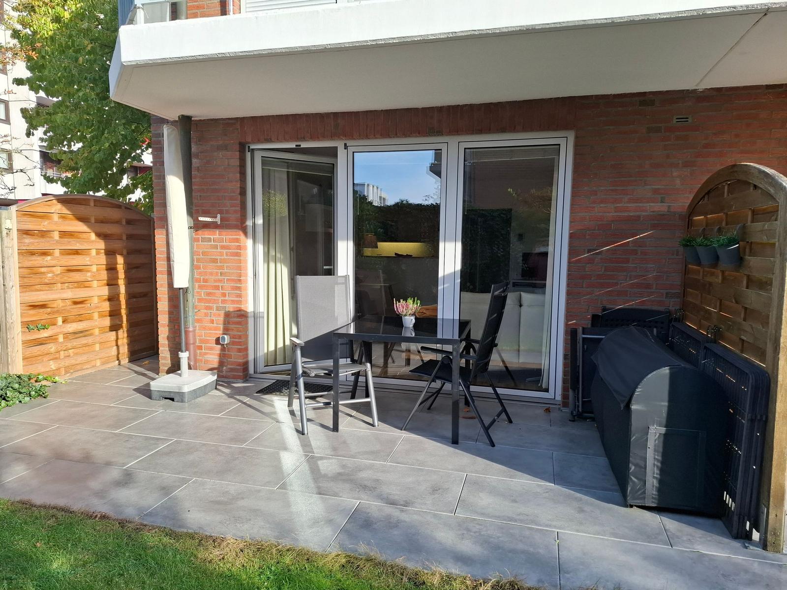 Terrasse mit Tisch, Stühlen und Grill. Glasfront führt ins Haus. Holzumrandung und Grasfläche.