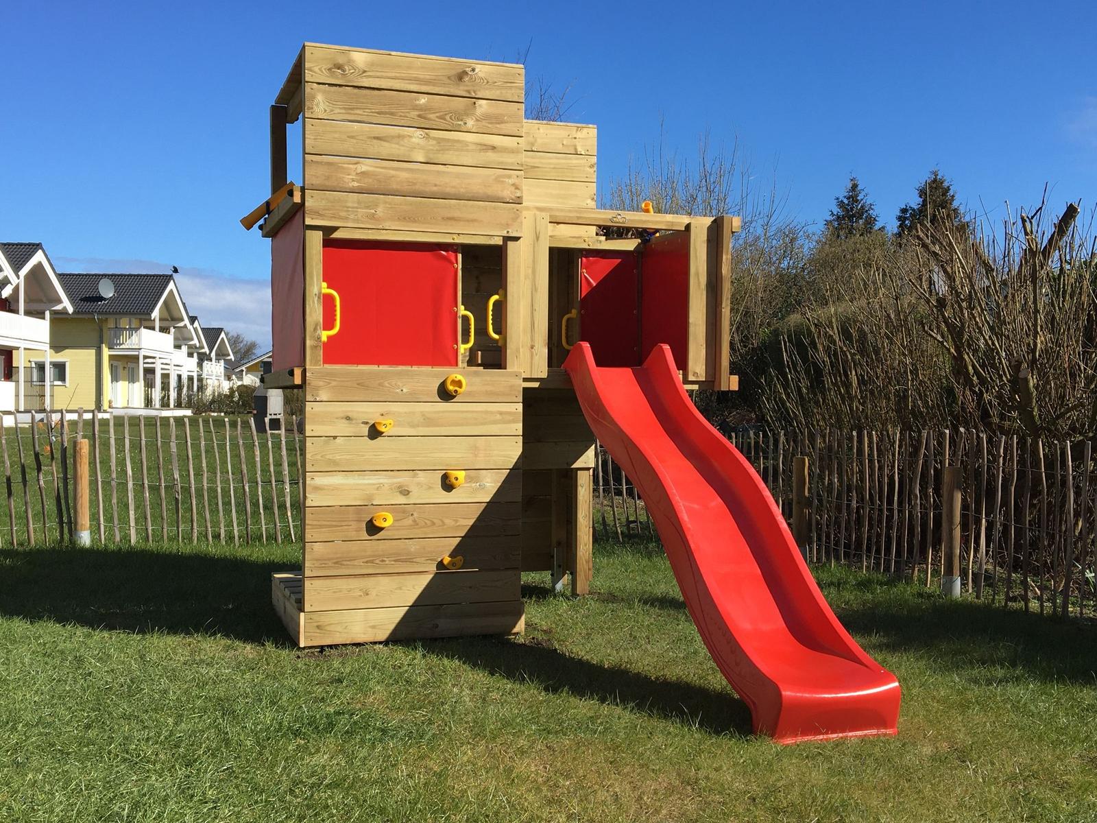 Holzspielhaus mit roter Rutsche und Kletterwand im Garten.
