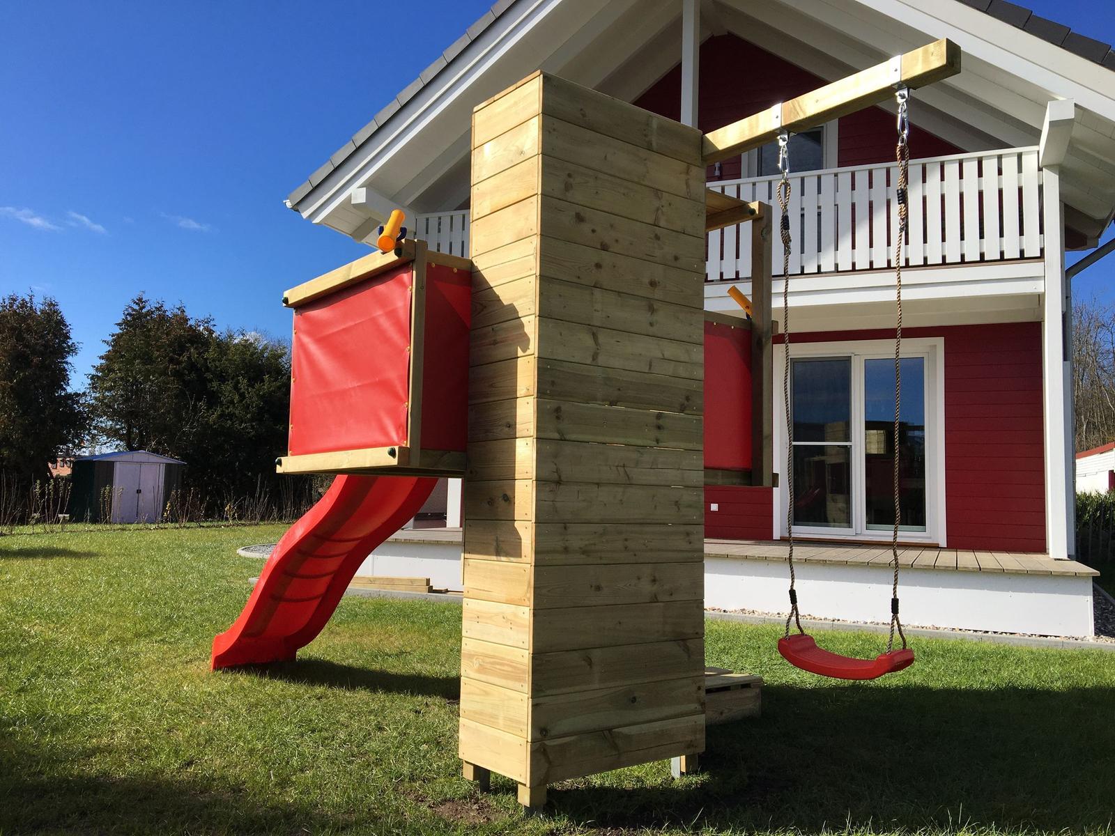 Holzspielplatz mit Rutsche und Schaukel vor einem roten Haus.