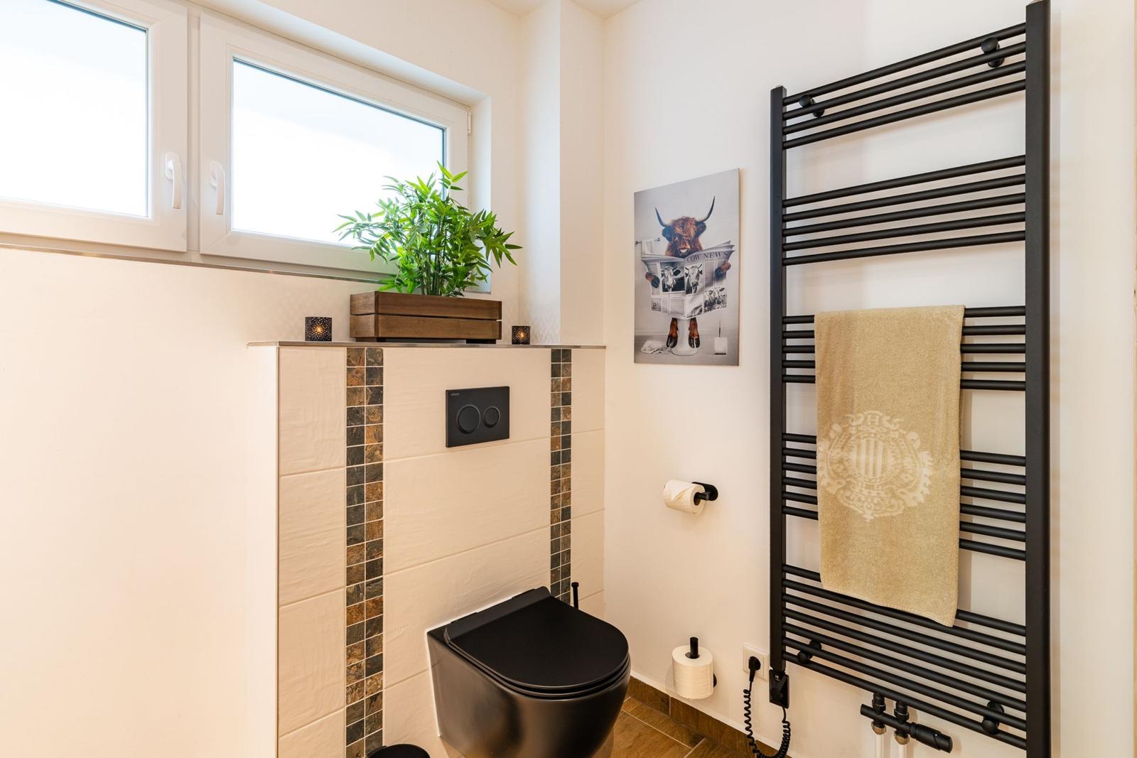 Badezimmer mit Toilette, Heizkörper und Fenster. Wandbild und Pflanze sichtbar.