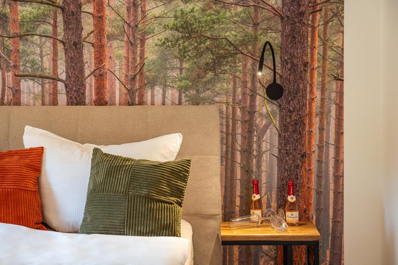 Schlafzimmer mit Waldtapete, Bett mit Kissen und Nachttisch mit Wein.