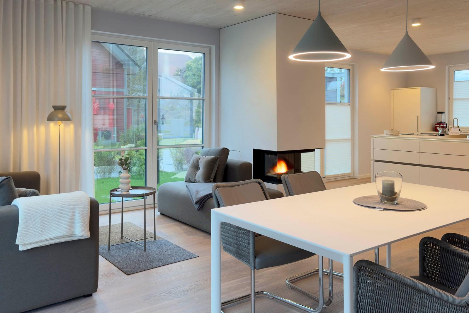 Moderne Wohnküche mit Esstisch, Sofa und Kaminofen. Große Fenster mit Blick auf Garten.