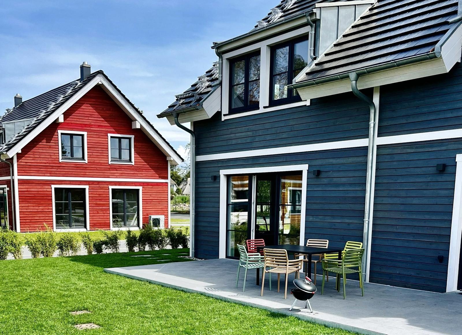 Ein blau-weißes Haus mit Terrasse und Grill. Neben einem roten Haus mit grünem Rasen.