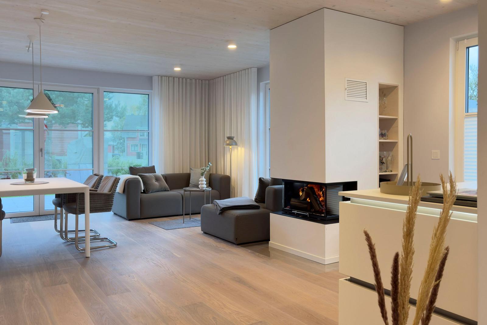 Wohnbereich mit Kamin, Sofa und Esstisch. Holzboden und große Fenster.