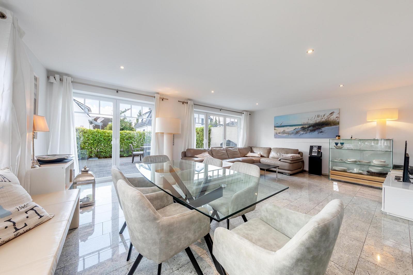 Ein Essbereich mit Glas-Tisch und Stühlen. Im Hintergrund ein Wohnbereich mit Sofa und großen Fenstern.