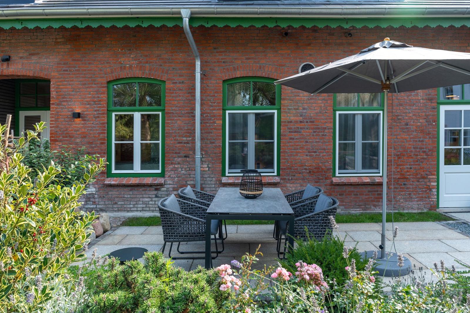 Red brick building with green window frames and garden furniture under a sun umbrella.