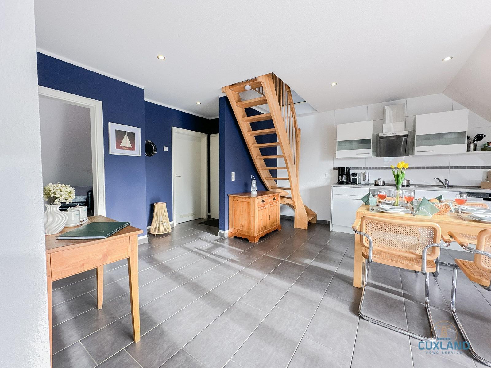 Küche mit Esstisch, Holztreppe und blauen Wänden. Wohnbereich mit Tisch und Blumen.