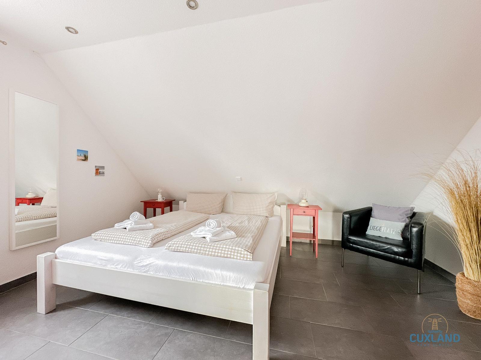 Schlafzimmer mit Doppelbett, Sessel und Spiegel an schräger Wand.