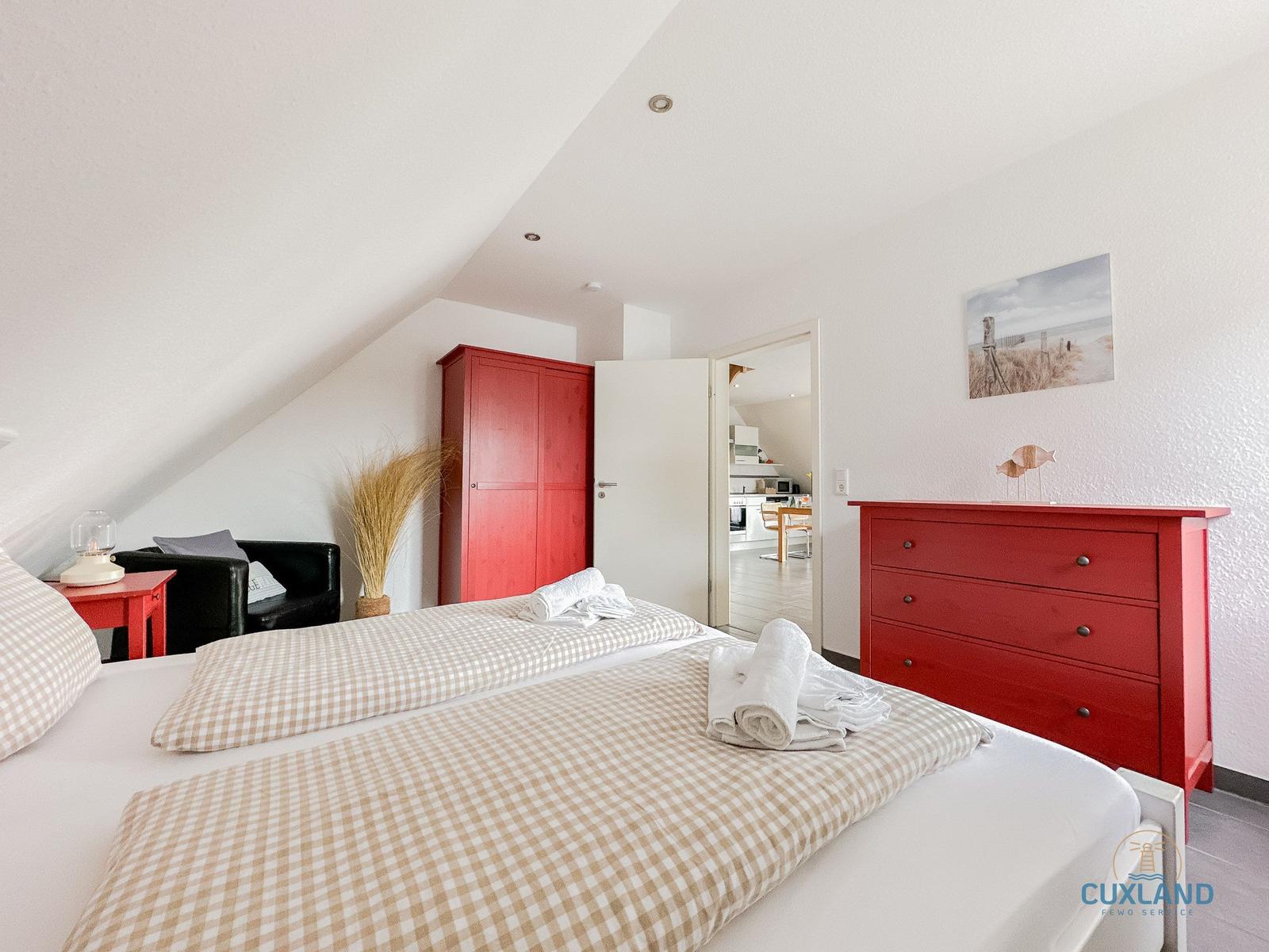 Schlafzimmer mit rotem Schrank und Kommode, Bett mit kariertem Bettzeug, Blick in die Küche.