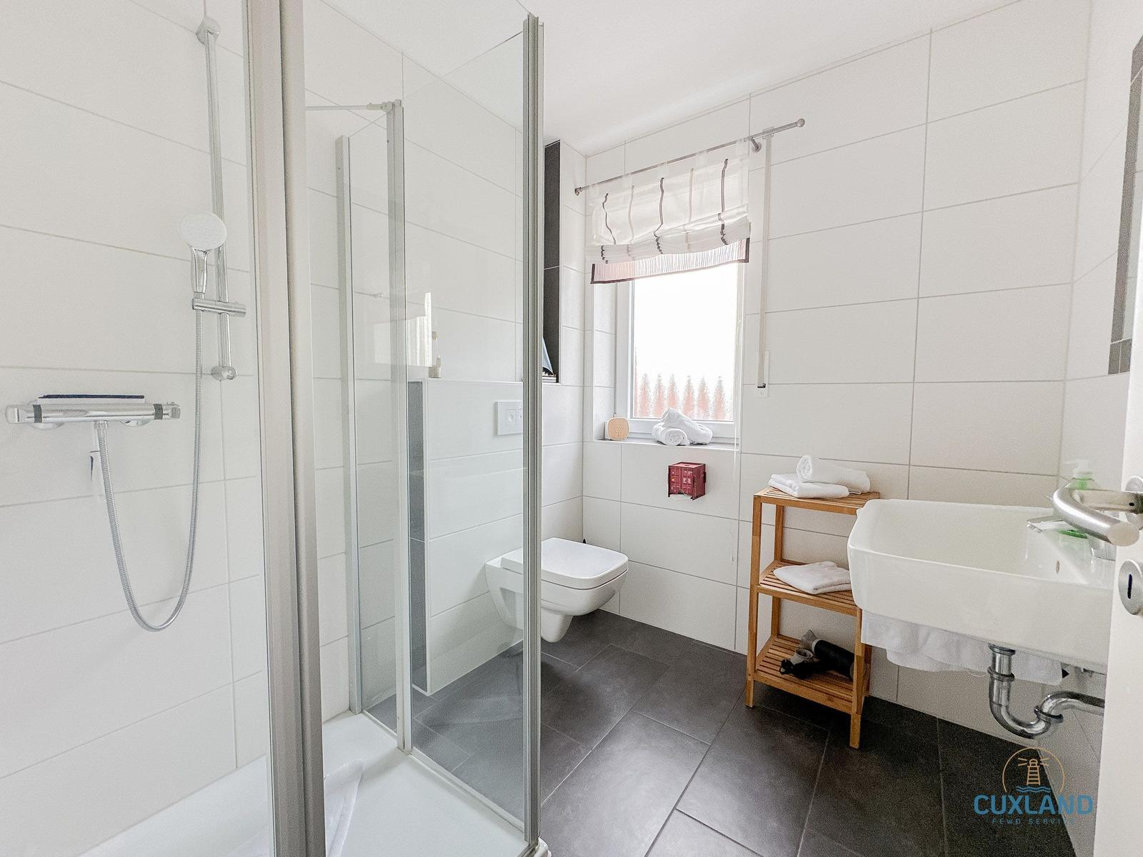 Badezimmer mit Dusche, Toilette und Waschbecken. Fenster mit Rollladen und Holzregal.