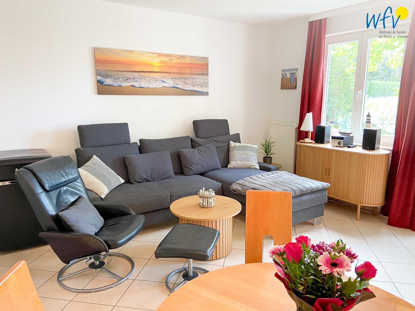 Wohnzimmer mit grauem Sofa, Ledersessel und Holztisch. Fenster mit roten Vorhängen.