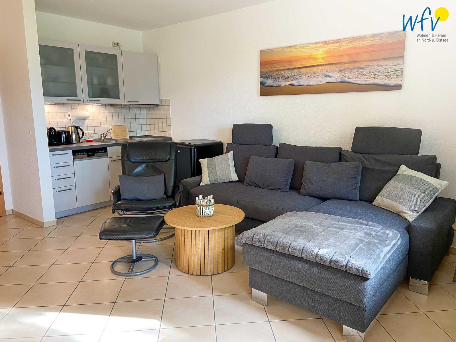 Wohnzimmer mit Sofa, Couchtisch und Küchenbereich. Wandbild zeigt Strand bei Sonnenuntergang.